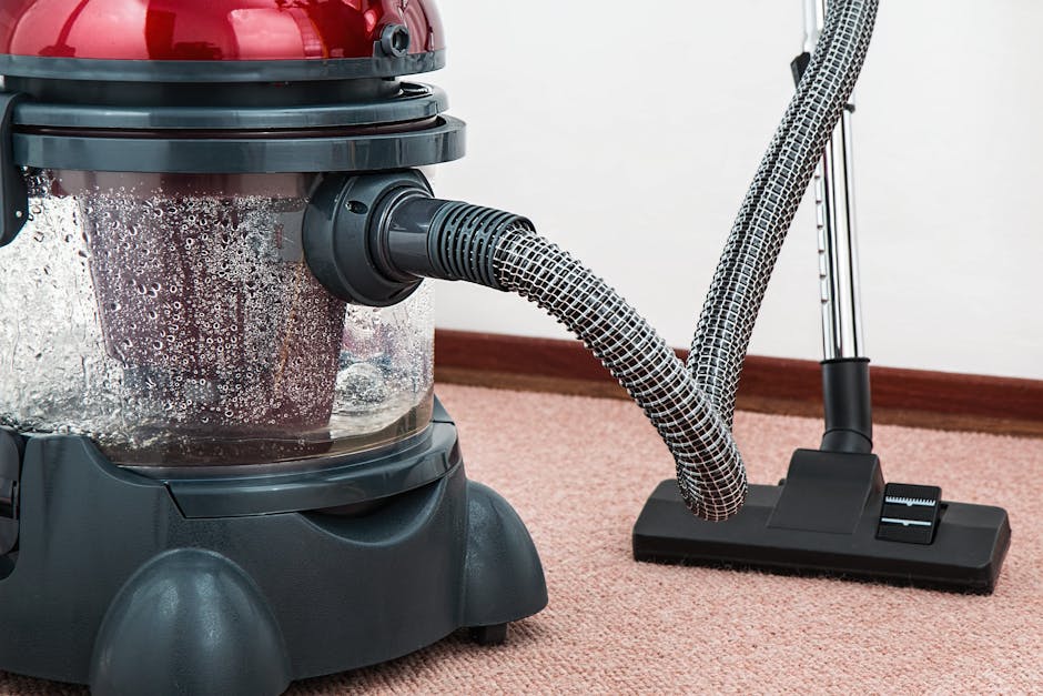 A woman kneeling on a patterned carpet in a living room, using a yellow and black vacuum cleaner for surface cleaning. The vacuum hose extends across the carpet, which appears clean and well-maintaine