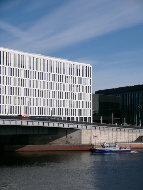 A view of modern commercial buildings along a riverfront, featuring large glass windows and sleek architectural designs with white and black facades. The scene includes a concrete walkway and a small blue boat sailing on the calm water below, under a partly cloudy sky. The area appears clean and well-maintained, suggesting professional surface cleaning and maintenance, relevant to commercial cleaning services offered by Carpet Cleaning Putney for Putney Bridge Riverside businesses.