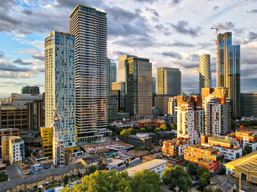 A panoramic view of a modern city skyline featuring numerous skyscrapers and high-rise buildings, including a tall, light blue glass tower with the 'Novotel' sign on it. The buildings are made of glass, steel, and concrete, reflecting the partly cloudy sky with patches of sunlight illuminating the structures. In the foreground, there are low-rise buildings and construction sites with visible cranes, surrounded by green trees and urban streets. The scene depicts contemporary architecture and urban development, emphasizing the structural variety and cleanliness of the cityscape as seen in natural daylight. The overall impression is of a vibrant, dynamic city environment with well-maintained surfaces and ongoing construction activity, highlighting the importance of surface cleaning and hygiene in urban spaces, relevant to residential and commercial cleaning services provided by Carpet Cleaning Putney.