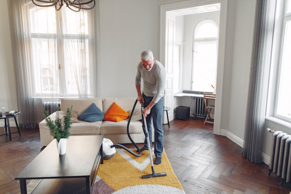 A man with gray hair wearing a light gray sweater and dark jeans is vacuuming a yellow geometric-patterned rug in a bright living room with ample natural light coming through large windows. The room features hardwood flooring, a white sofa with colorful cushions, a small black side table with a potted green plant, and sheer curtains. The windows have white trim and are positioned on white walls, with a radiator visible beneath the window on the right. The cleaning activity involves surface cleaning and deep cleaning, emphasizing hygiene and maintenance, and is performed by Carpet Cleaning Putney as part of their domestic cleaning services.