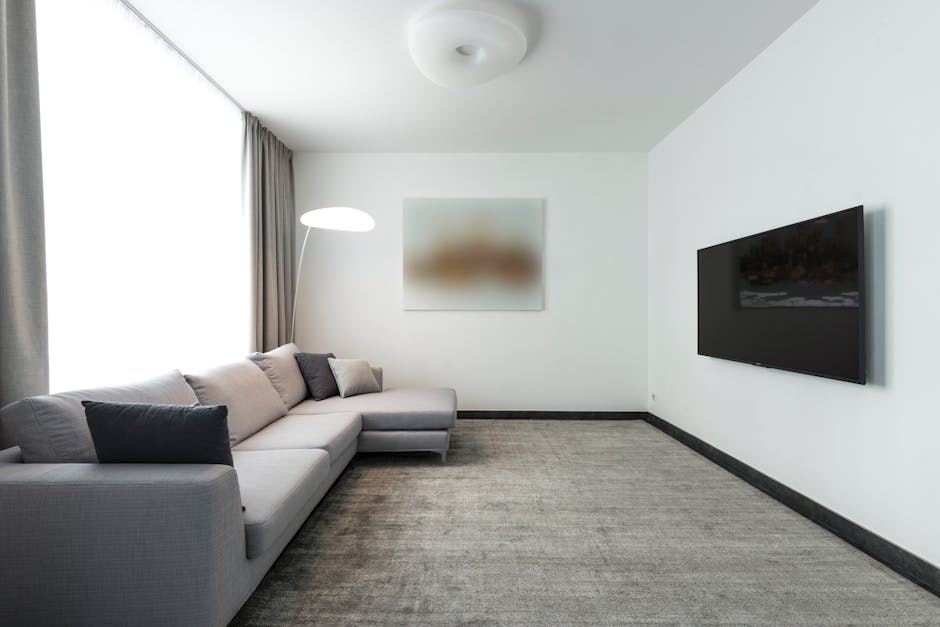 A minimalist living room with white walls and a large window covered by light gray curtains, allowing natural light to illuminate the space. A light gray fabric sofa with multiple matching and darker accent cushions is positioned against the left wall. A tall, arched floor lamp with a white shade stands beside the sofa, providing additional lighting. On the right wall, a large flat-screen television is mounted, with a clean, reflective screen. The floor is covered with a gray textured carpet that appears clean and well-maintained. An abstract, blurred artwork hangs centrally on the wall above the sofa, contributing to the modern and tidy ambience of the room, which is part of a professional cleaning service by Carpet Cleaning Putney, highlighted by the neatness and overall hygiene of the space, suitable for interior sanitisation and domestic deep cleaning.