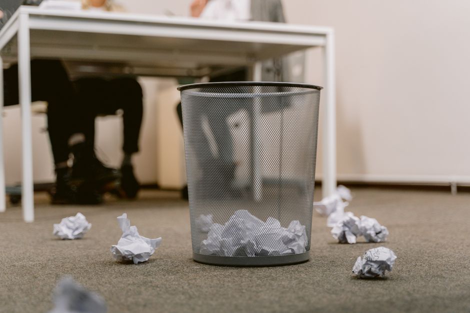An office scene featuring a metal mesh trash bin placed on a beige carpeted floor, filled with crumpled sheets of paper. Several crumpled papers are scattered around the bin. In the background, there is a white desk with a person seated behind it, partially visible, with only their legs and black shoes showing, and a white wall. The lighting appears to be natural or standard indoor lighting, highlighting the cleanliness of the carpet and the organised workspace. Carpet Cleaning Putney offers surface cleaning and deep cleaning services focused on maintaining hygiene and cleanliness in office environments, aligning with Wandsworth Council rules for proper carpet waste disposal in Putney.