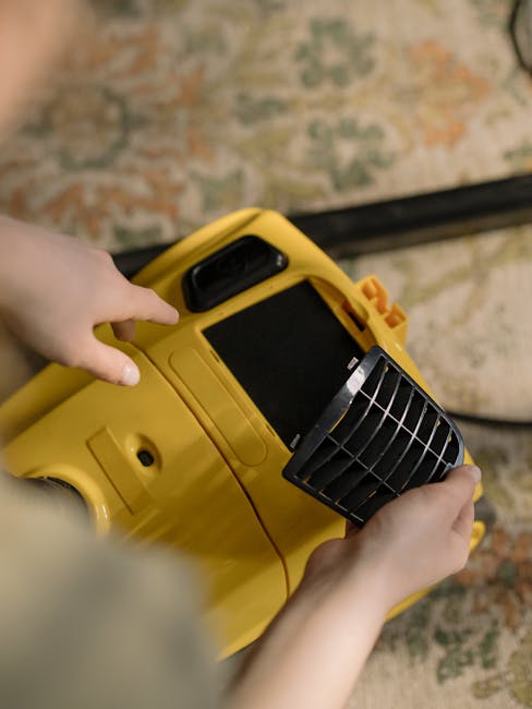 A person holding a yellow industrial carpet cleaning machine, with their left hand gripping the handle and their right hand adjusting a black grille vent, positioned over a patterned carpet in a well-lit room. The carpet features intricate designs in muted tones of beige, orange, and green, and appears clean and well-maintained. The cleaning device has a large black control panel and a sturdy design, indicating professional deep cleaning equipment used by Carpet Cleaning Putney as part of surface cleaning and sanitisation services. The background shows a portion of the room's carpeted floor, emphasizing the focus on professional domestic or commercial carpet cleaning.